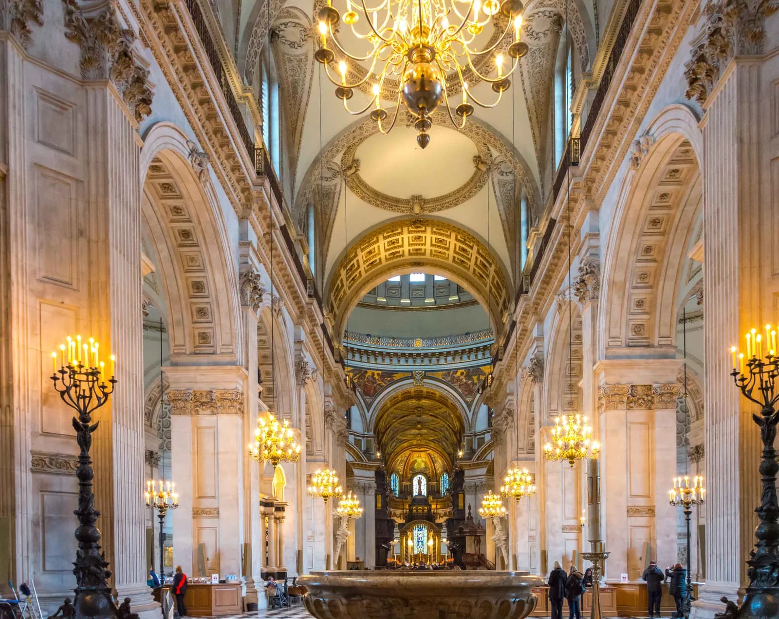 Ornate chandeliers illuminate the grand interior of a cathedral with high arched ceilings, columns, and a large baptismal font in the foreground.