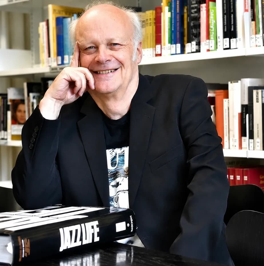 A smiling man with white hair wearing a black jacket and t-shirt, resting his chin on his hand, sits at a table in a library with a large book titled 'JAZZ LIFE' in front of him.